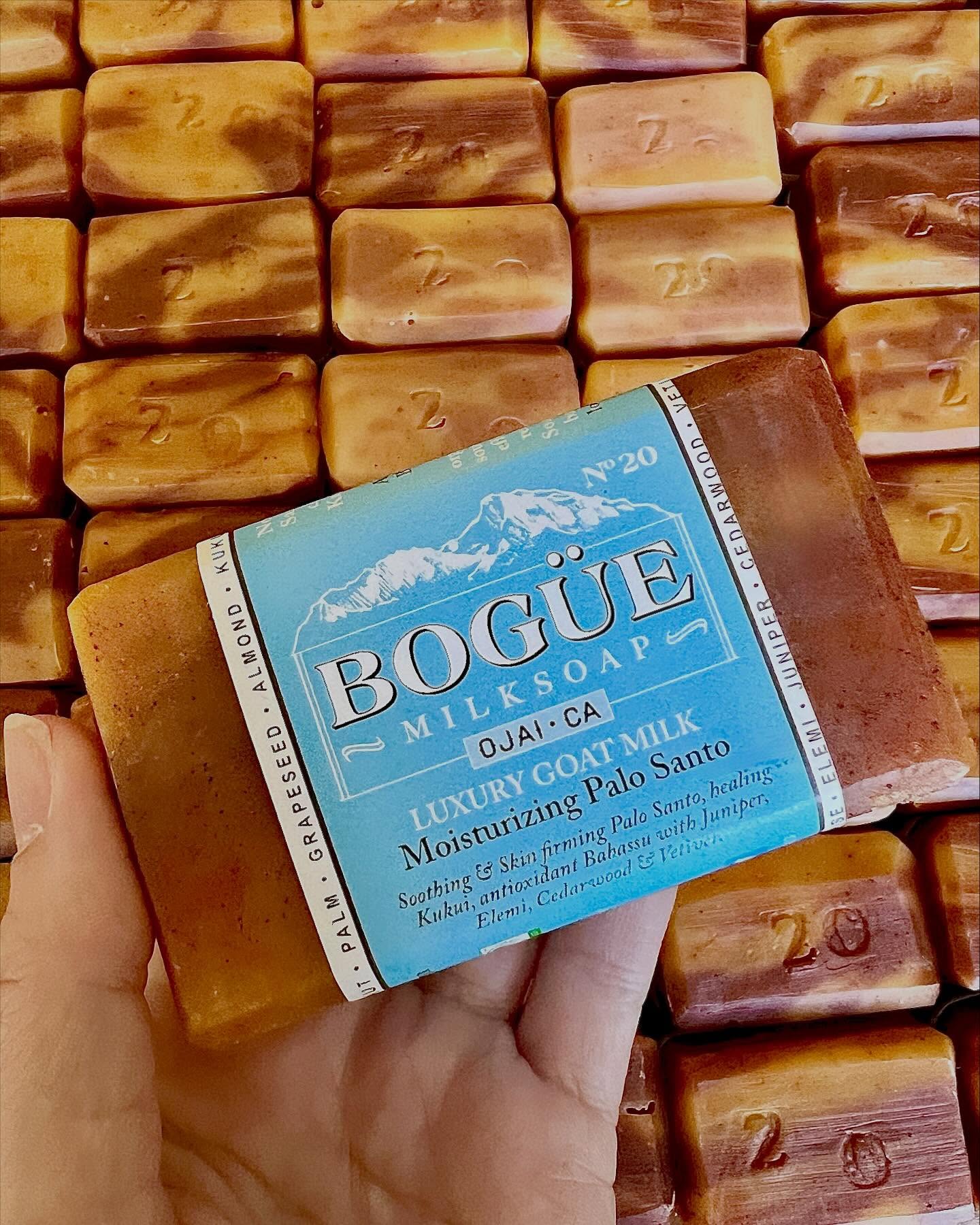 Hand holding Bogue Milk Soap bar labeled “Moisturizing Palo Santo” over a background of freshly made goat milk soap bars in Ojai, California