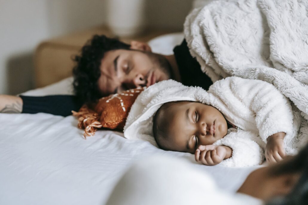 Multiethnic father and newborn baby lying and sleeping together on comfortable bed near crop unrecognizable woman in daylight